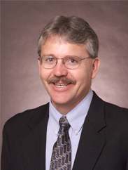 Wade Brorsen smiling in a black suit and grey tie in front of a brown background.