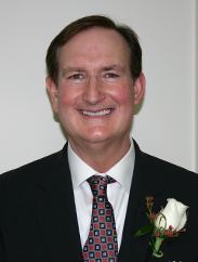 Clay Pollard smiling in a black suit and tie against a white backdrop.