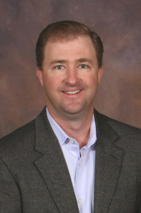 Cody Catalena in a dark grey suit, smiling against a brown background