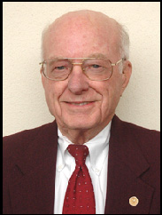 Don Kaspar smiling in a black suit and red tie against a cream background