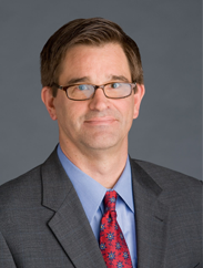 Doug Duncan smiling in a dark grey suit and red tie against a grey background