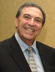 Edward Smith smiling in a black suit and grey tie against a yellow background.