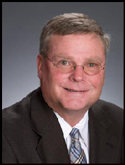 Joseph Swinbank smiling in a  black suit and grey tie against a grey backdrop