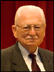 Portrait of Ivan Schmedemann smiling in a grey suit against a dark red.