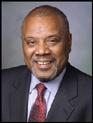 Frederick McClure smiling in a black suit and red tie against a blue-grey backdrop