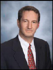 Gary Gunter smiling in a black suit and red tie against a blue background.