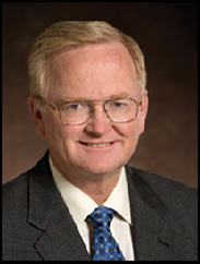 James Dodson smiling in a black suit and blue tie against a dark brown background.