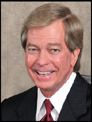 Bill Carter smiling in a black suit and red tie against a light brown backdrop.