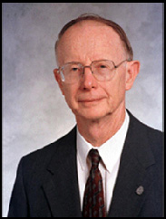 Carl G. Anderson, in a black suit and tie, smiling in front of a grey scale background.