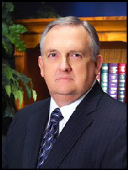 Jimmy Wright straight faced wearing a black suit and blue striped tie in front of a bookcase