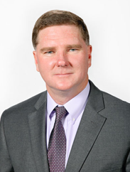 John Miller smiling in a grey suit and purple tie against a white backdrop