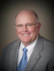 Kirk Cleere smiling in a grey suit and blue tie against a grey background.