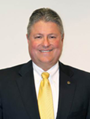 Mark Humphrey smiling in a black suit and yellow tie in front of a white backdrop