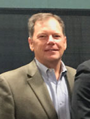 Mark Miller in a grey suit smiling against a grey background