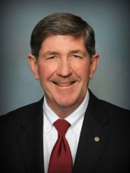 Paul Bracher in a black suit and red tie, smiling in front of a blue lit background.