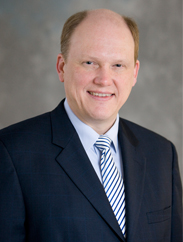 Troy Thompson smiling in a black tie and blue striped tie against a grey backdrop