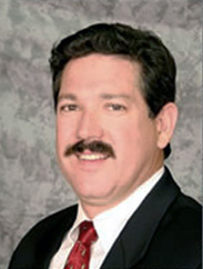 Raul Hinojosa smiling in a black suit and red  tie in front of a grey background.