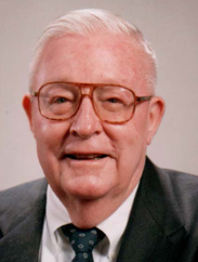 Mack Gray smiling in a black suit and tie in front of a grey background.