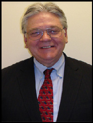 Stanley Johnson smiling in a black suit and red tie against a cream background