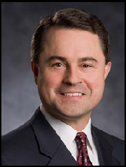 Tod Staples smiling in a black suit and red tie against a black backdrop