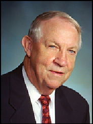 Tom Smith smiling in a black suit and red tie against a blue backdrop