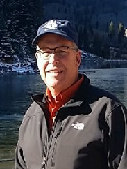 Nick bamert in front of a lake, wearing a blue cap and grey jacket, smiling.