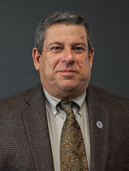 John Malazzo smiling in a dark grey suit and green/gold tie against a grey background