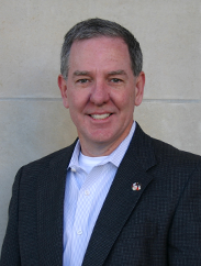 Rob Hall smiling in a black suit in front of a white stone background.