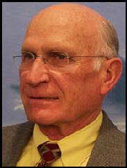 Portrait of Walter Richburg in a grey suit and yellow button up in front of a blue backdrop