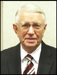 Zay Gilbreath smiling in a black suit and red/white tie against a cream background.