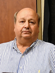 John Giesenschlag in a striped blue and white button up in front of brown and black background.