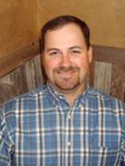 Justin Bierschwale in blue flannel button down, smiling in front of wood paneling background.