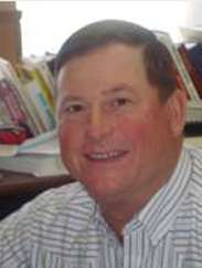 Paul Bierschwale smiling in a striped white and blue collared shirt in front of a stack of books. 