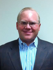 William Corrigan smiling in a black suit and white/blue striped button down against a blue backdrop.