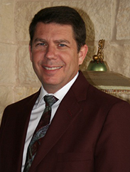 Mike Martin smiling in a maroon suit and black tie in front of a light stone background