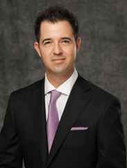 Casey Oldham smiling in a black suit and purple tie against a dark grey backdrop