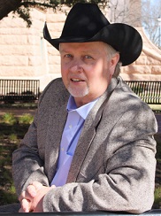 Stan Bevers in a grey coat and black cowboy hat leans against a post in the outdoors.